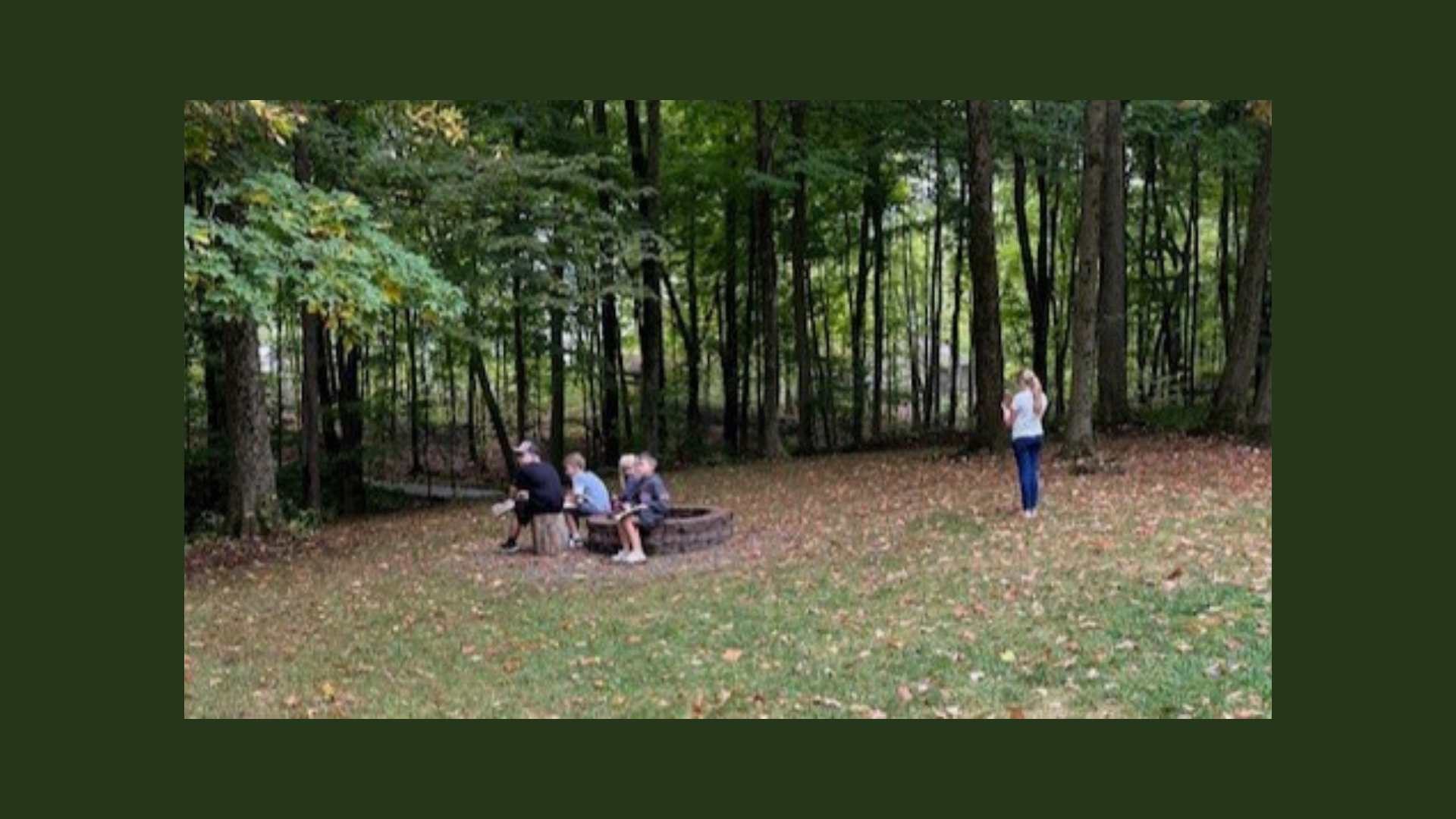 a small group of kids sitting on the edge of a stone fire ring during the day and another kid near the trees