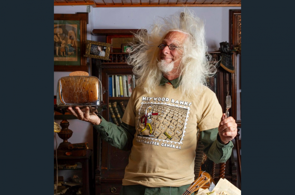 Haywood Banks with long white hair blowing around his face, holding a toaster and a fork, and wearing a t-shirt that says Haywood Banks, toastmaster general