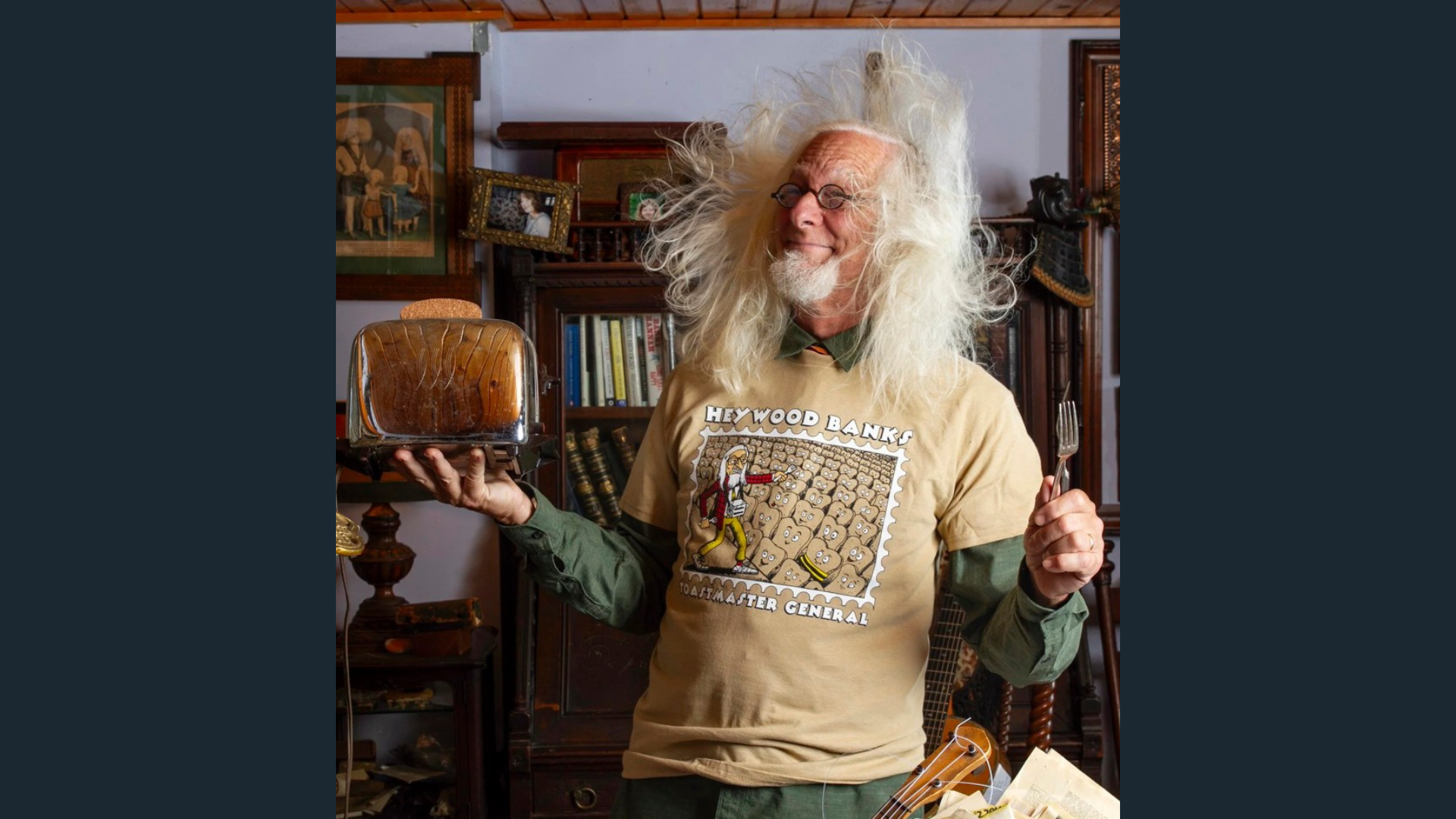 Haywood Banks with long white hair blowing around his face, holding a toaster and a fork, and wearing a t-shirt that says Haywood Banks, toastmaster general