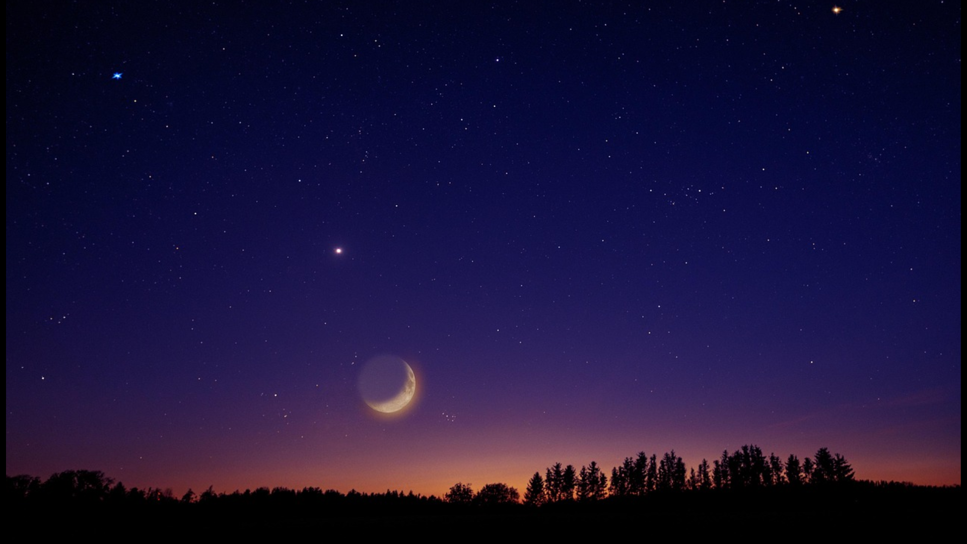 A sliver of the moon in a starlit sky at night with a tree line in silhouette